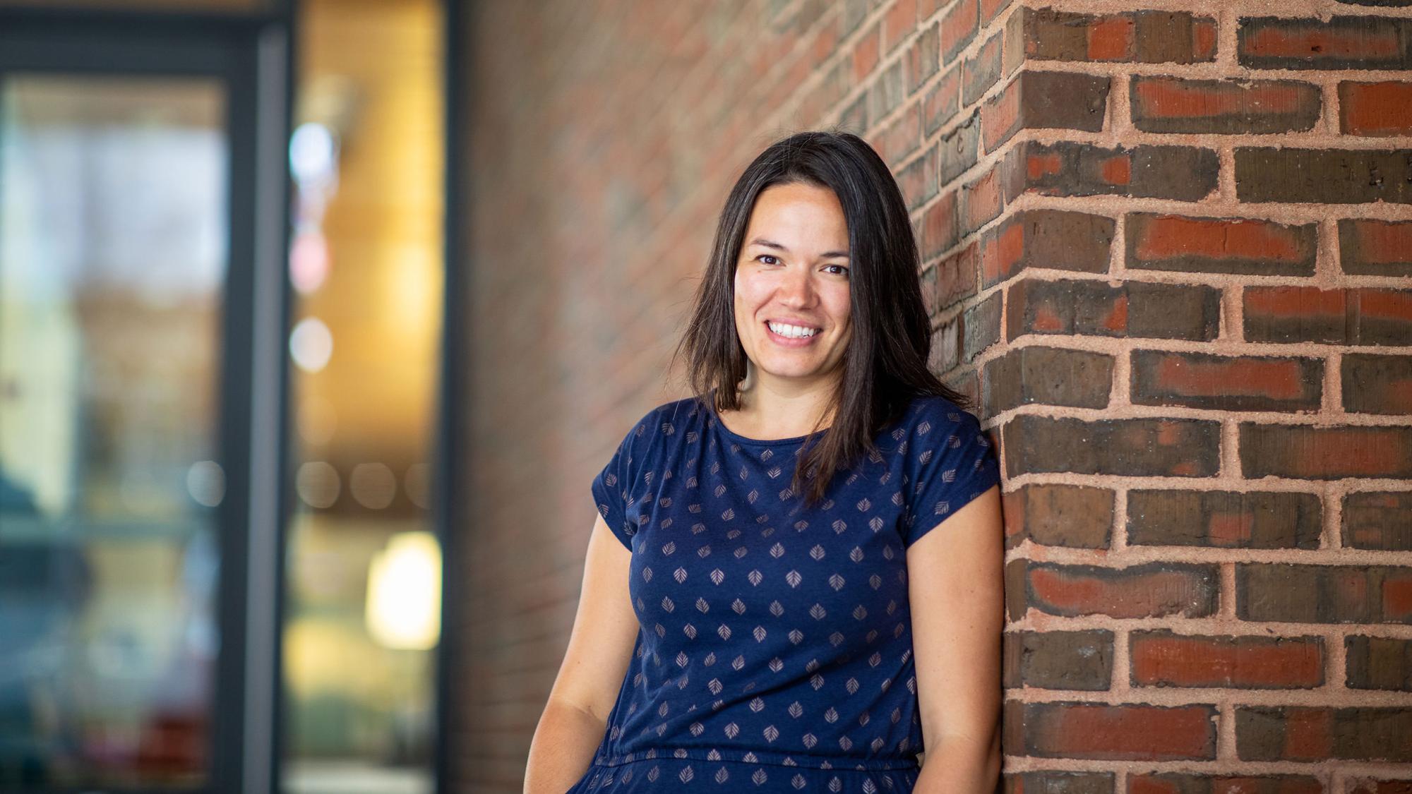 Elizabeth Linos is smiling for the camera in a short sleeve blue shirt standing against a brick wall