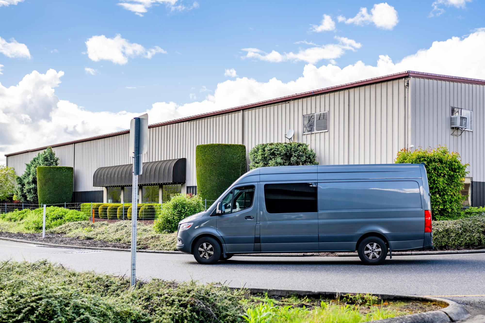 A grey van driving on a paved street in front of a white industrial building.