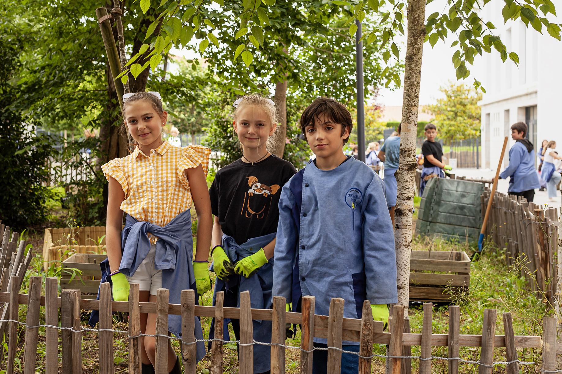 Two young girls and a boy stand with gloves on in a garden