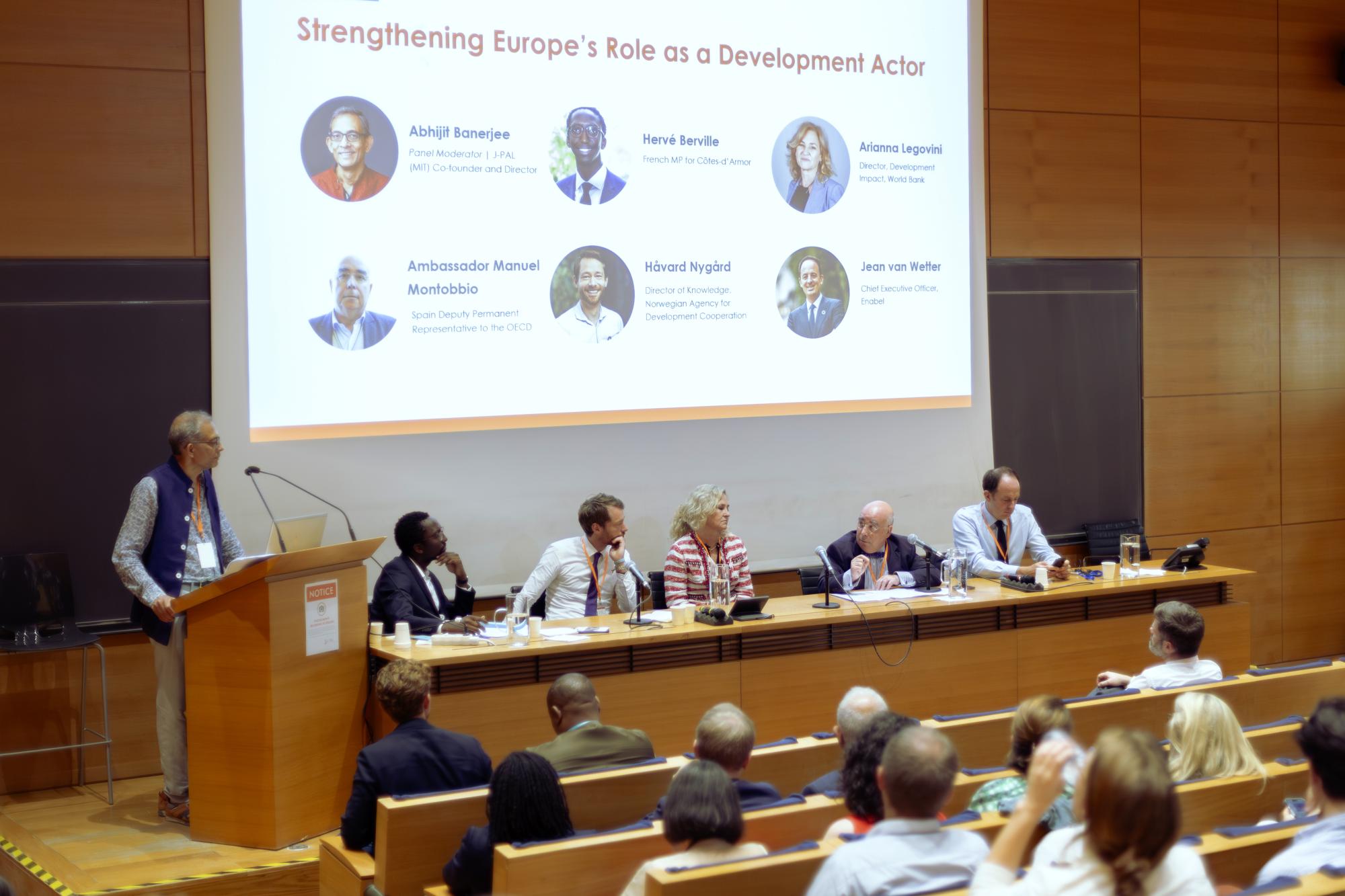 Abhijit Banerjee behind a podium moderating a panel of five speakers who are seated. 