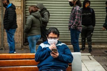 A man sits on a bench wearing a mask and looking at his phone. There is a line of people behind him.