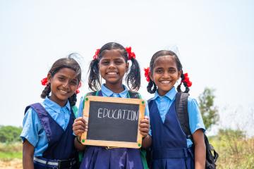 Three school girls stand with a chalkboard sign that reads "Education."