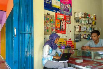 A man stands behind a counter that displays phone cards in a small shop. A woman sits in front of the counter typing on a laptop. 