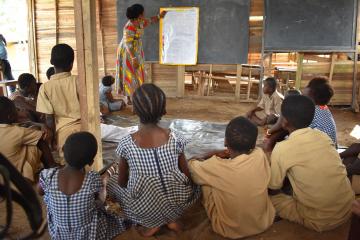 Children sit in a ring around a teacher, who is demonstrating something on a blackboard