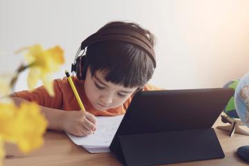 A young boy wearing headphones writes on a piece of paper in front of a laptop