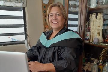 A woman wearing academic dress sits in front of a laptop and smiles at the photographer.