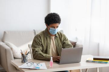 A man wearing a mask sits at a desk typing on a computer.