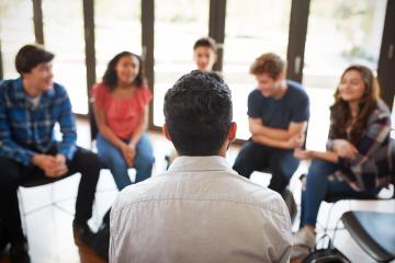 A circle of people sitting in chairs face a man sitting on a chair in front of them