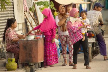 Three women, two holding babies, buy food from a vendor seated at a table.