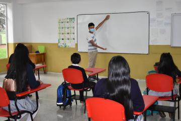 A young man stands at a whiteboard in front of a class of students, all wearing masks