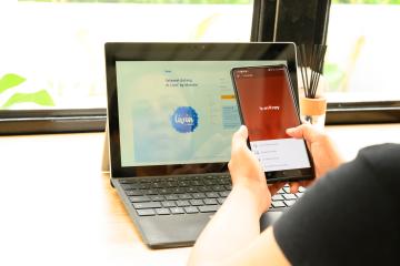 A person uses an e-money app on a phone while sitting in front of a laptop. 