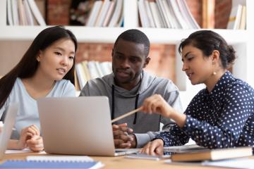 Three people of different ethnicities sit in front of a laptop and discuss.