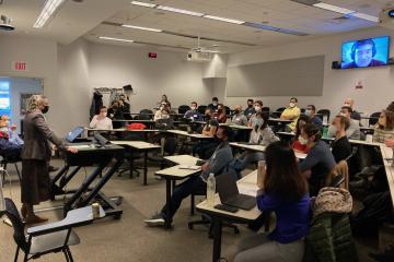 A lecturer stands in front of a screen, facing a room filled with students seated at desks.