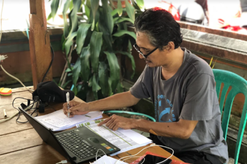 A woman writes down data on a notepad while consulting a laptop in Indonesia