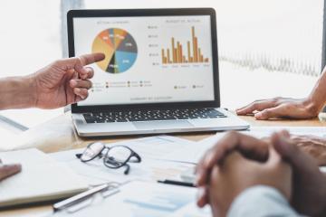 People refer to various charts on a laptop during a meeting.