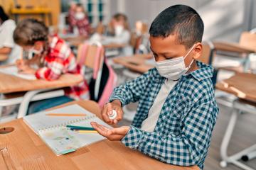 A student sits at their desk applying hand sanitizer and wearing a face mask.