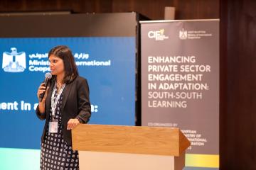 Neha Sharma stands at a podium holding a microphone, with a poster in the background that reads "Enhancing Private Sector Engagement in Adaptation: South to South Learning"