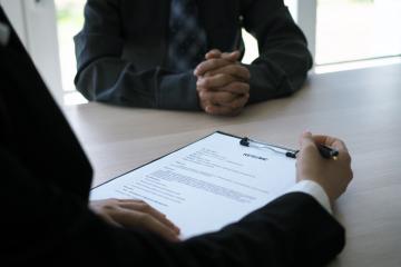 Two individuals at a table, one with a pen and clipboard with paper with text "Resume"