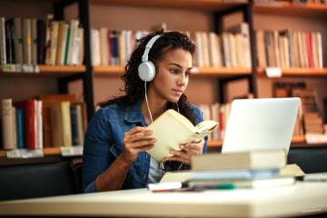 A woman wearing headphones and holding a book studies in a library and looks at a laptop.