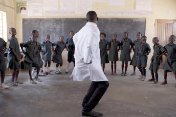 A group of children in a classroom encircle a person dancing in front of them.