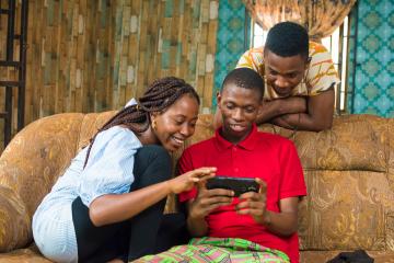 A young woman and two men smile while watching a show on a phone. 