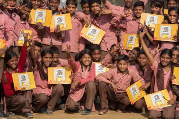 A large group of smiling schoolchildren in uniform proudly hold up “Taaron Ki Toli” learning kits, posing together outdoors with hands raised in excitement.