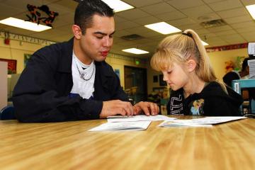 a man wearing a white t-shirt and black sweater points at a book on a desk, alongside a girl with blonde hair pulled up in a ponytail. 