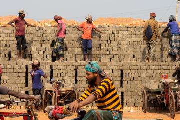 Bangladeshi traditional kiln workers stack bricks and converse with other workers in the background. In the foreground, other workers transport brick loads with bicycles. 