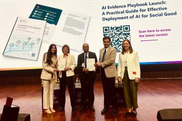 Five men and women stand together on a stage facing the audience. One man is displaying a book to the audience. The background reads, "Launch of the AI Evidence Playbook."