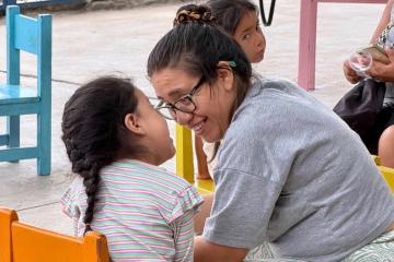 A woman with brown hair and glasses in a grey shirt smiles at a young girl with brown hair in a braid and white shirt. They both are seated at a table indoors.